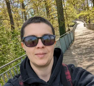 Selfie of a smiling short haired woman in a light forest wearing sunglasses.